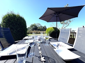 Een eettafel buiten, gedekt met glazen, borden en bestek, onder een grote parasol, met uitzicht op een tuin en bomen.
