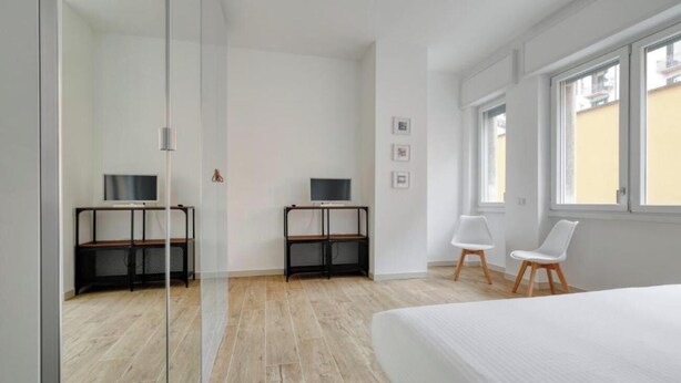 A modern bedroom with a large bed, two desks with computers, a chair, and a window with a view of buildings.