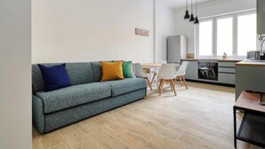 A modern living room with a grey sofa, a dining table, and chairs. There is a kitchen area with a stove and cabinets.