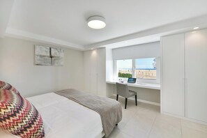A bedroom with a bed, a desk with a laptop, a window with a view, and a wardrobe.