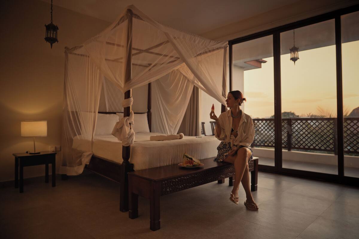 A bedroom with a four-poster bed, a nightstand, a bedside table, and a large window offering a view of the outdoors.