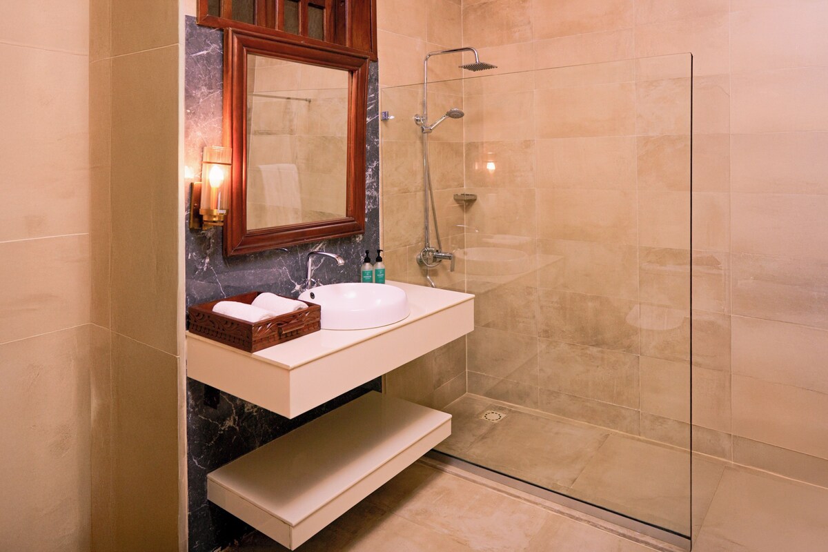Un baño con un lavabo blanco, tocador de madera, cabina de ducha con mampara de vidrio y paredes con azulejos color beige.