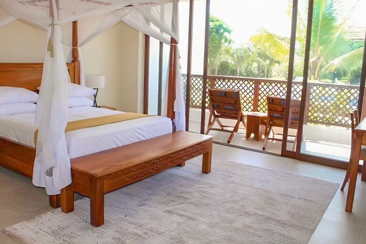 A bedroom with a canopy bed, a wooden bench, a balcony with a view of palm trees.