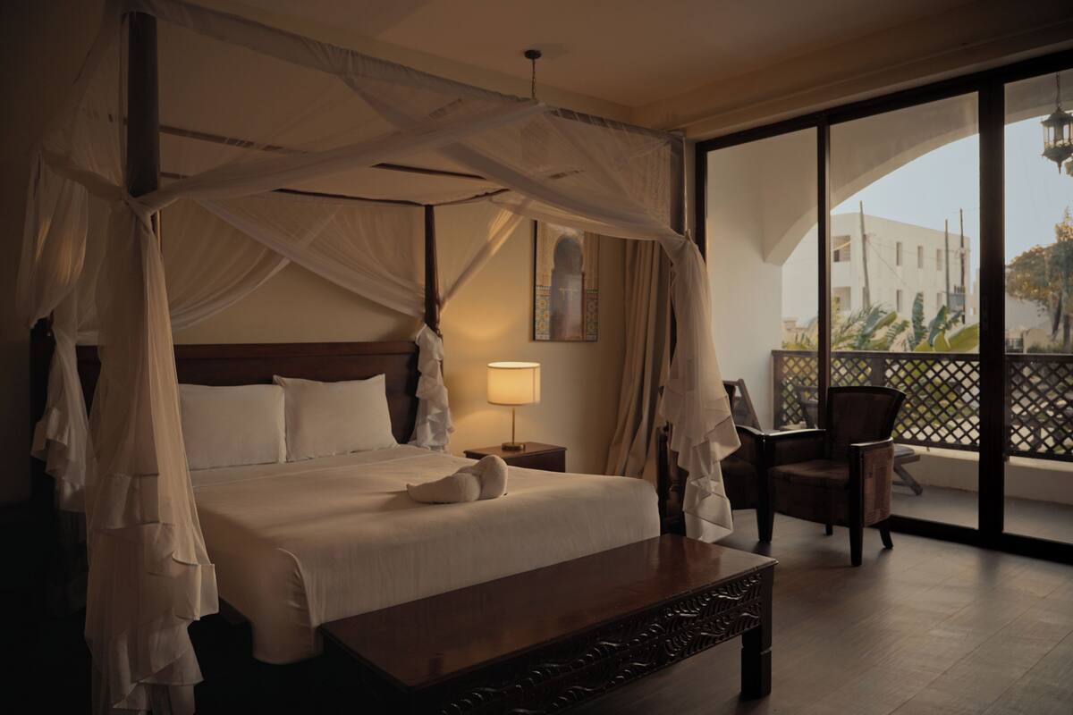 A four-poster bed with a canopy, a wooden nightstand, a chair, and a balcony with a view of buildings.