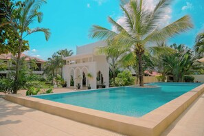Piscina com um edifício branco ao fundo, rodeada por palmeiras e vegetação.
