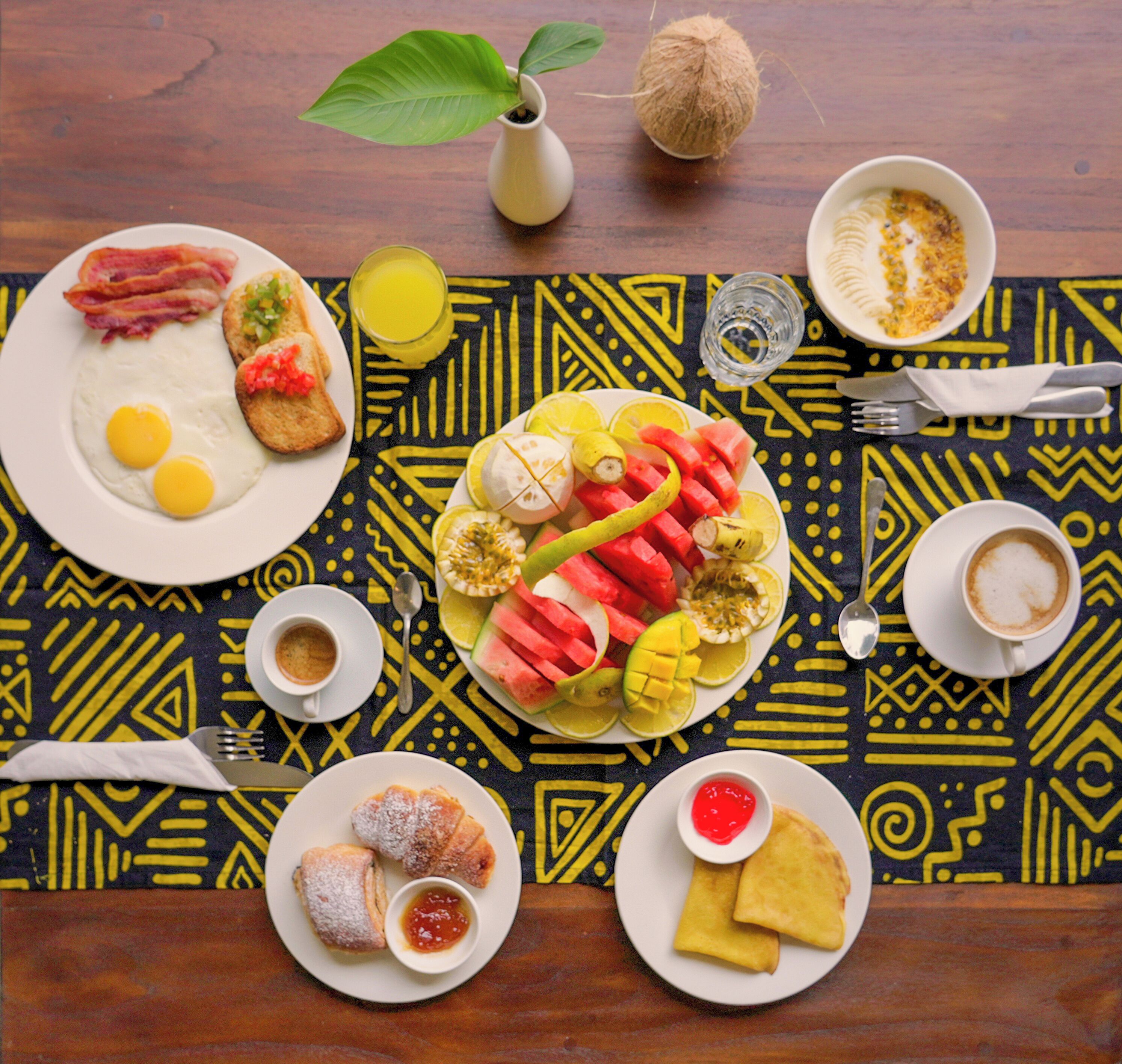 A breakfast table with plates of eggs, bacon, toast, fruit, pastries, and coffee.