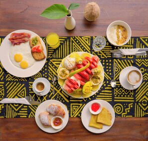 Mesa de café da manhã com pratos de ovos, bacon, torradas, frutas, doces e café.