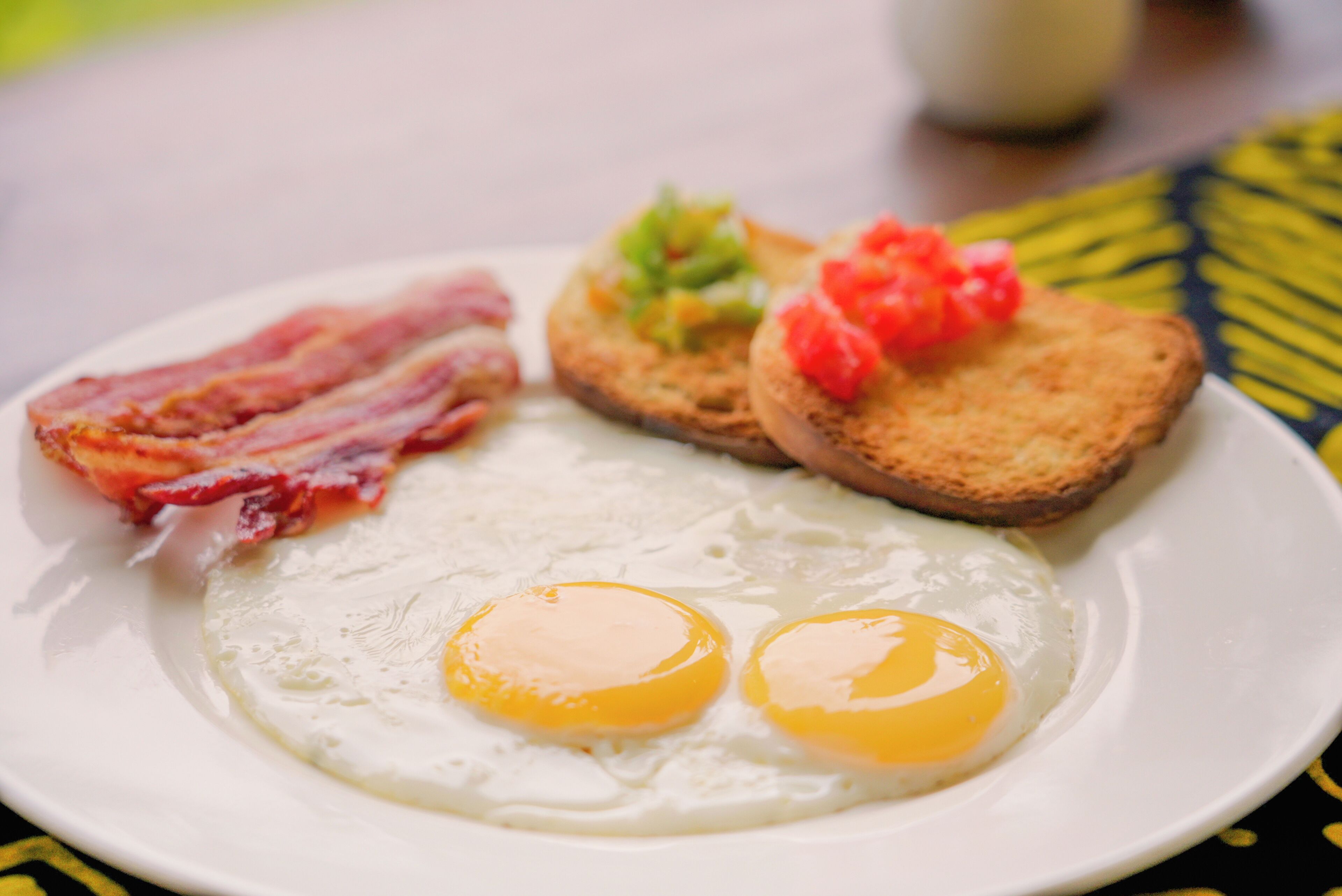 Un piatto da colazione con due uova fritte, bacon, pane tostato con pomodoro e guarnizioni verdi.
