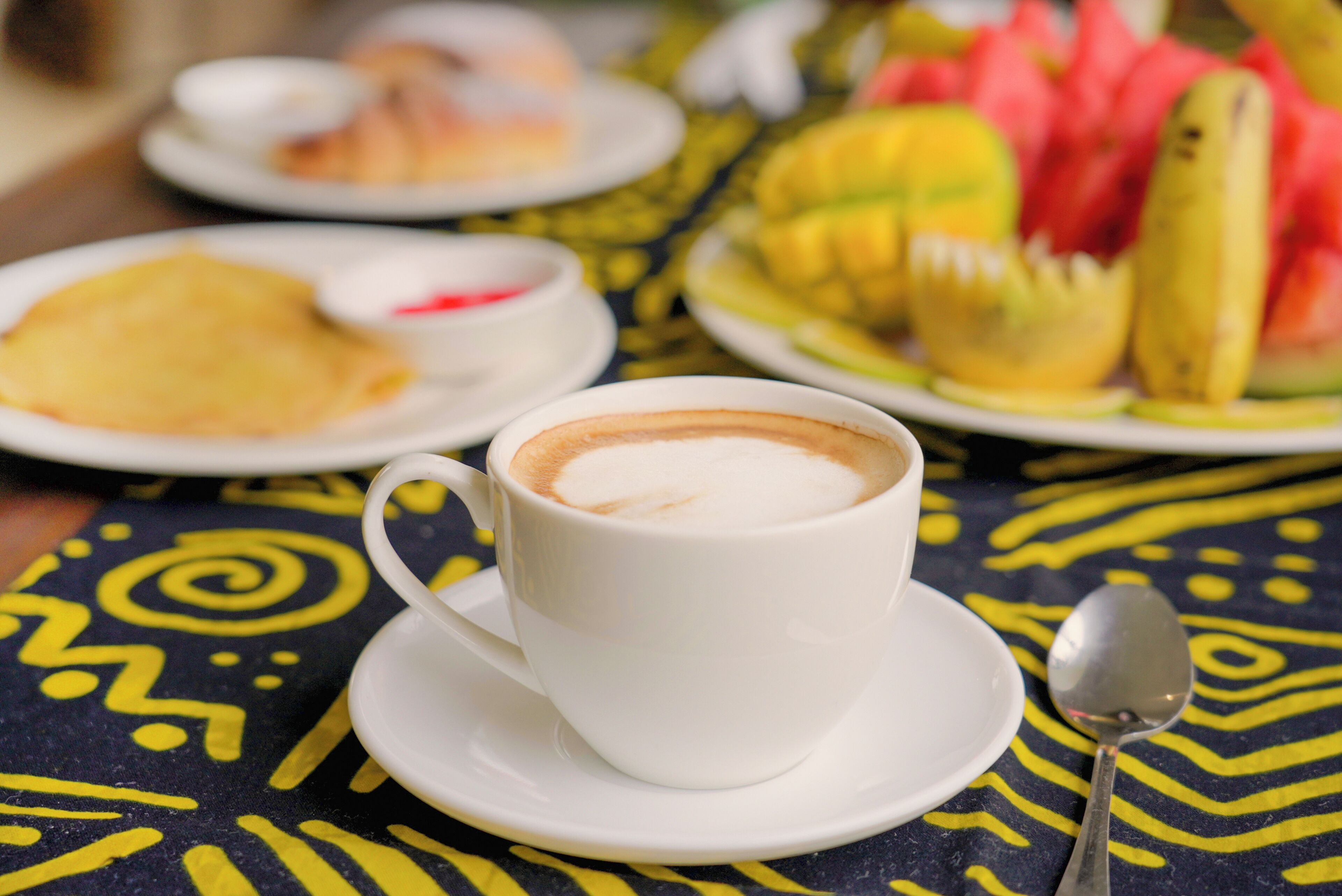 A cup of coffee on a saucer with a spoon beside it, plates of food, and a patterned tablecloth.