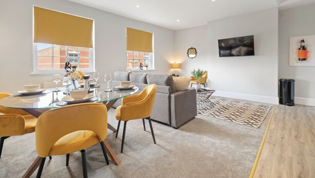 Un salon moderne avec une table à manger en verre, des chaises jaunes, un canapé gris et un tapis à motifs.