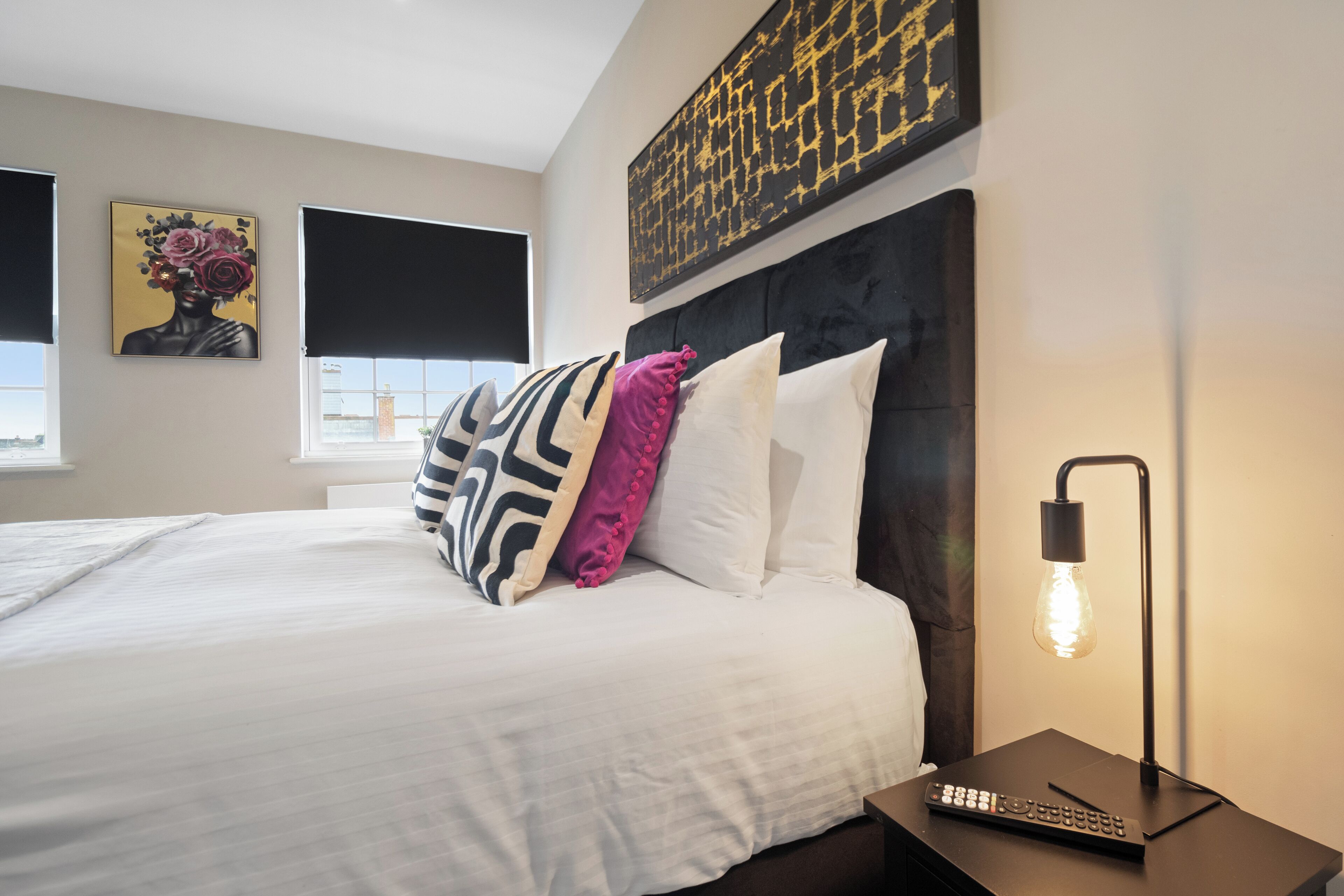 A neatly made bed with white linens and patterned pillows, a modern headboard, a wall-mounted artwork, and a bedside table with a lamp and remote control.