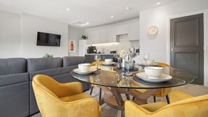 Un salon moderne avec un canapé gris, des fauteuils jaunes et une table à manger en verre, dressée pour quatre personnes.
