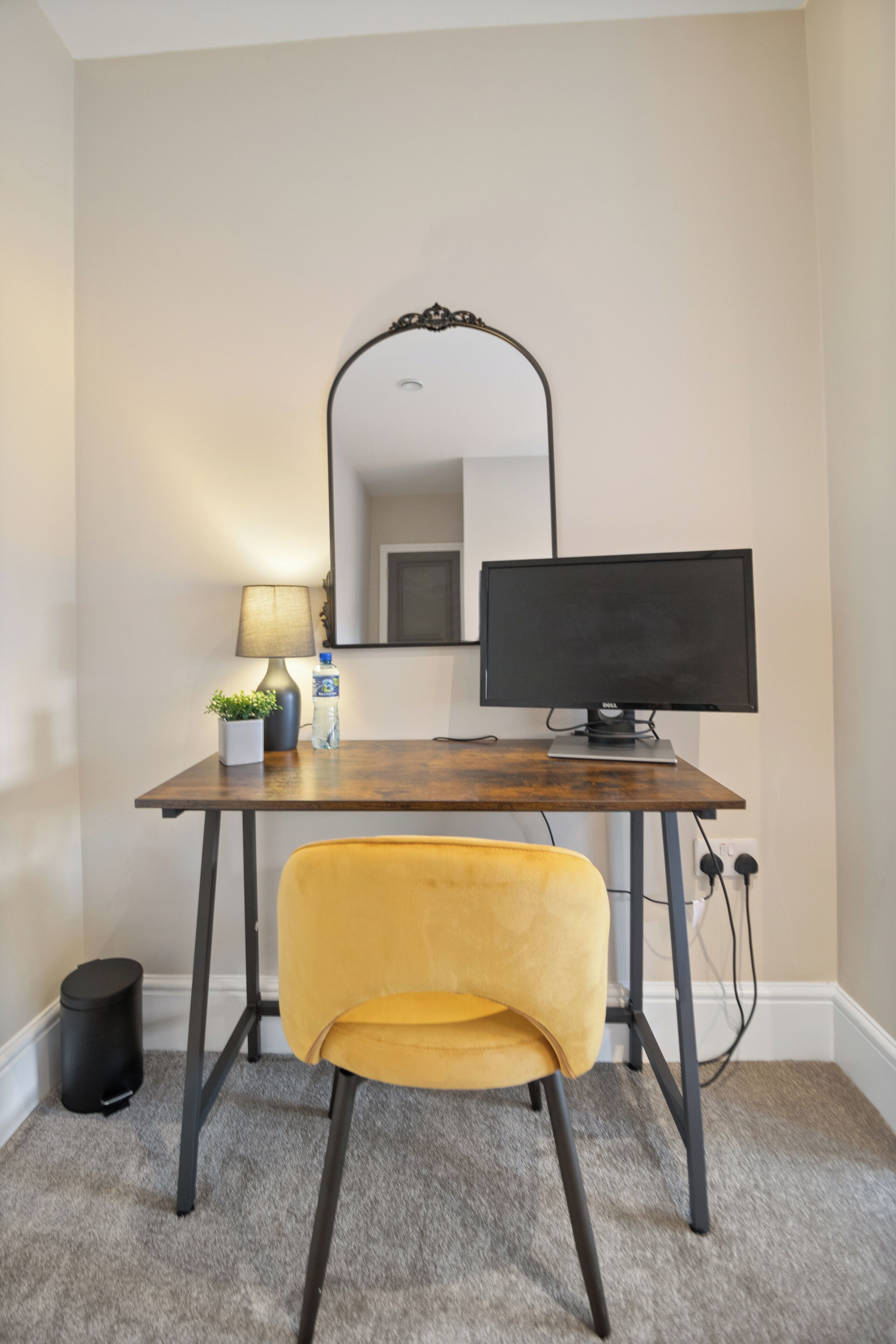 A small office space with a desk, a computer monitor, a lamp, a chair, and a mirror on the wall.