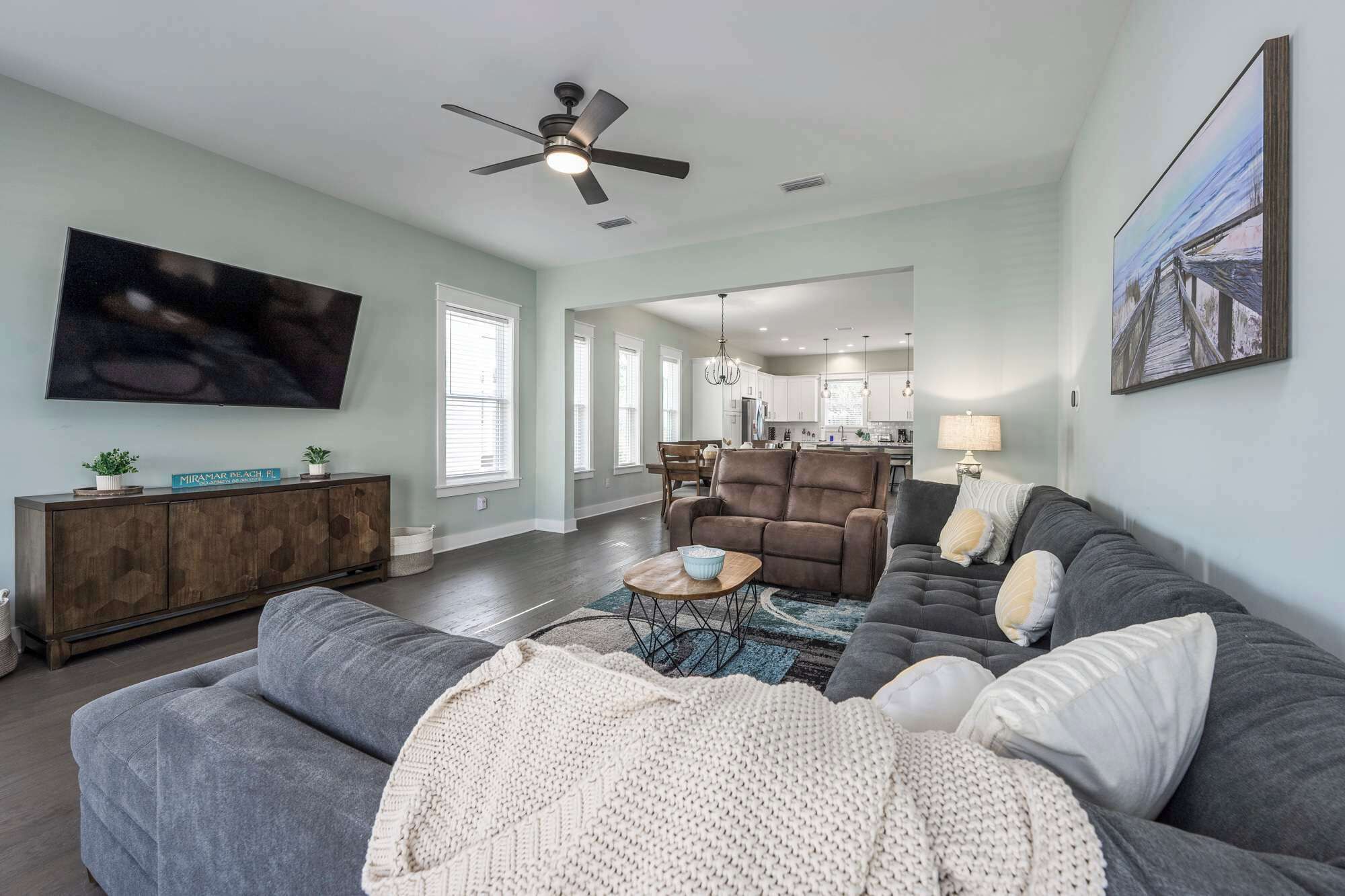 A spacious living room with a grey sofa, a brown recliner, and a TV mounted on the wall. There is a ceiling fan, a dining area with a table and chairs, and a kitchen in the background.