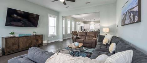 A spacious living room with a grey sofa, a brown recliner, and a TV mounted on the wall. There is a ceiling fan, a dining area with a table and chairs, and a kitchen in the background.