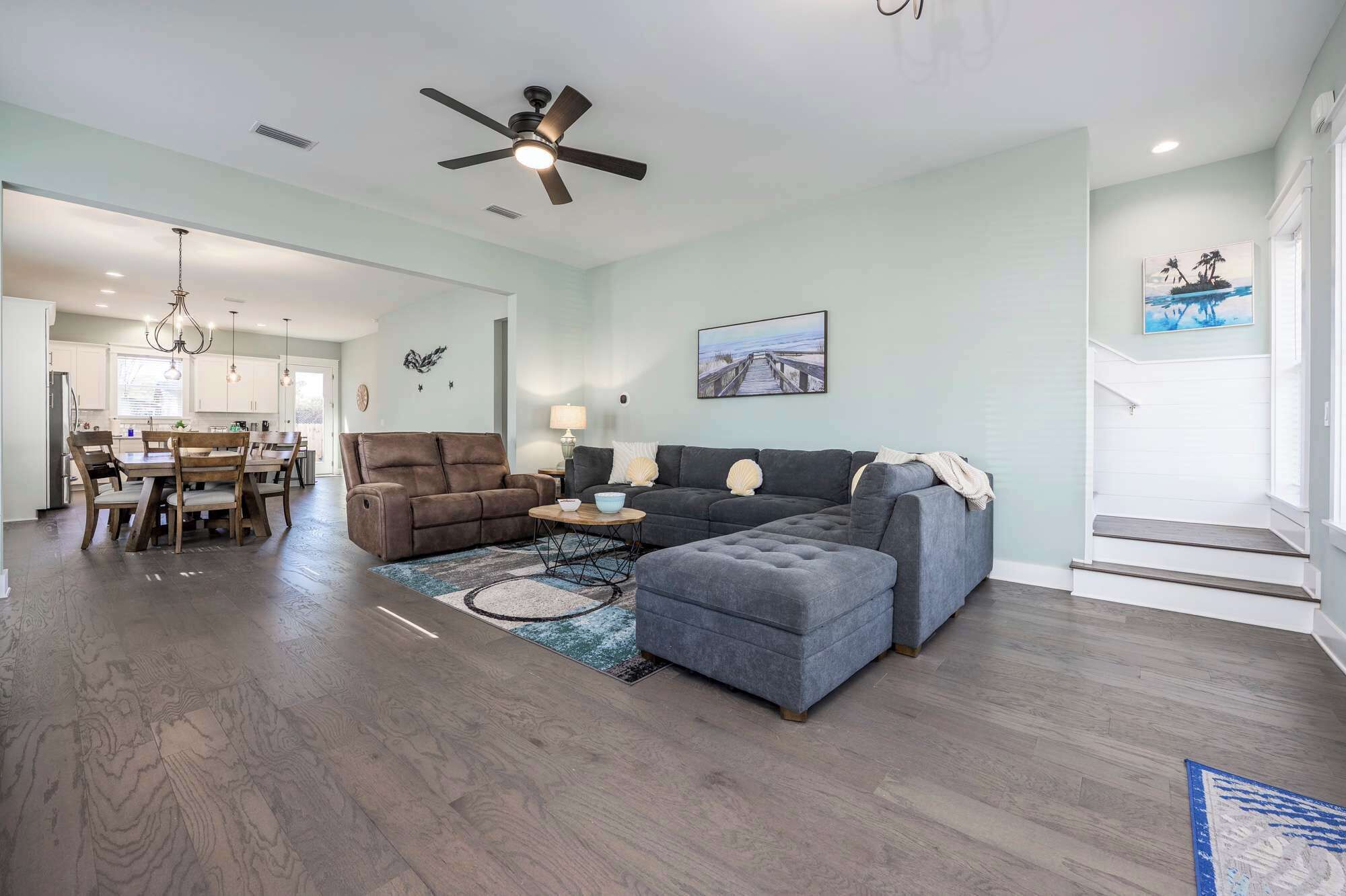 A spacious living room with a dark grey sectional sofa, a brown recliner, and a grey ottoman. A dining area with a wooden table and chairs is visible in the background.