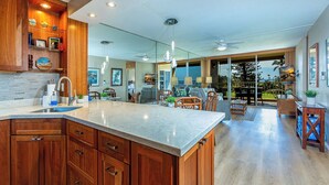 A modern kitchen with wooden cabinets, a marble countertop, and a large island. There is a dining area with a view of the outdoors.