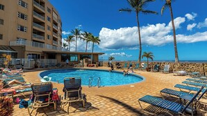 A pool area with lounge chairs, palm trees, and a building with balconies.