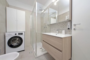 A modern bathroom with a washing machine, a glass shower enclosure, and a wooden vanity with a mirror. - YCH Bagnera Suites (Rome)