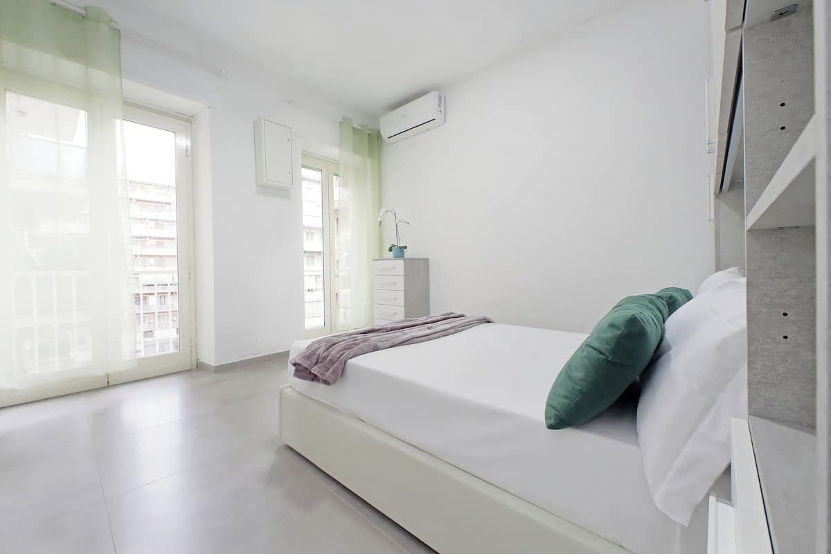 A bedroom with a bed, a window with a view, and an air conditioning unit.