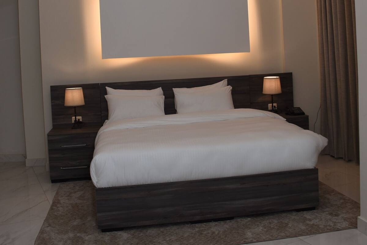 A modern bedroom with a large bed, dark headboard, two bedside lamps, and a recessed ceiling light.