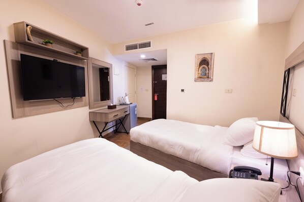 A hotel room with two beds, a desk, a TV, a lamp, and a small plant. - Sukoon Hotel Amman (Amman)