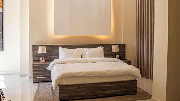 A modern bedroom with a large bed, a nightstand, a desk, and a large window with curtains. - Sukoon Hotel Amman (Amman)