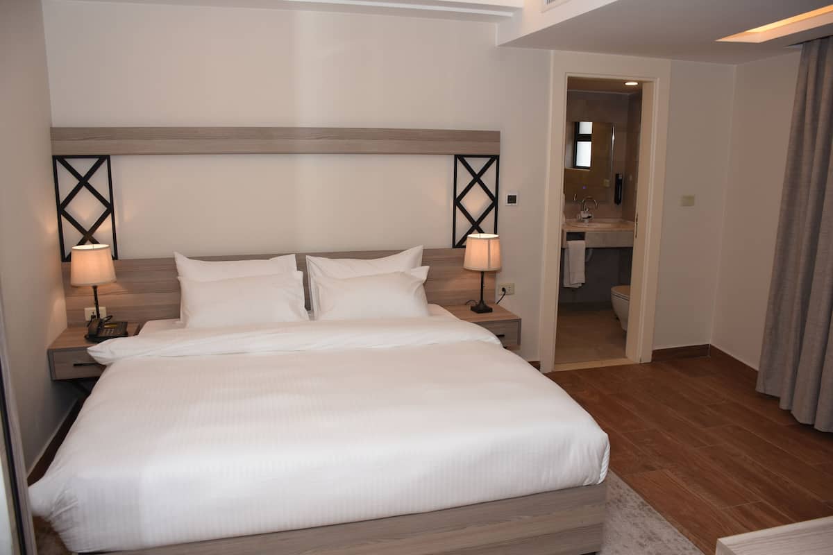 A hotel room with a large bed, two bedside tables with lamps, a wooden headboard, and a bathroom visible through an open door.