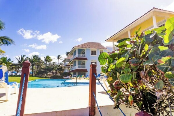 A pool area with lounge chairs, a large building with a red roof, and a tropical plant in the foreground. - K-2 Luxury Beachfront (Cabarete-Sabaneta)