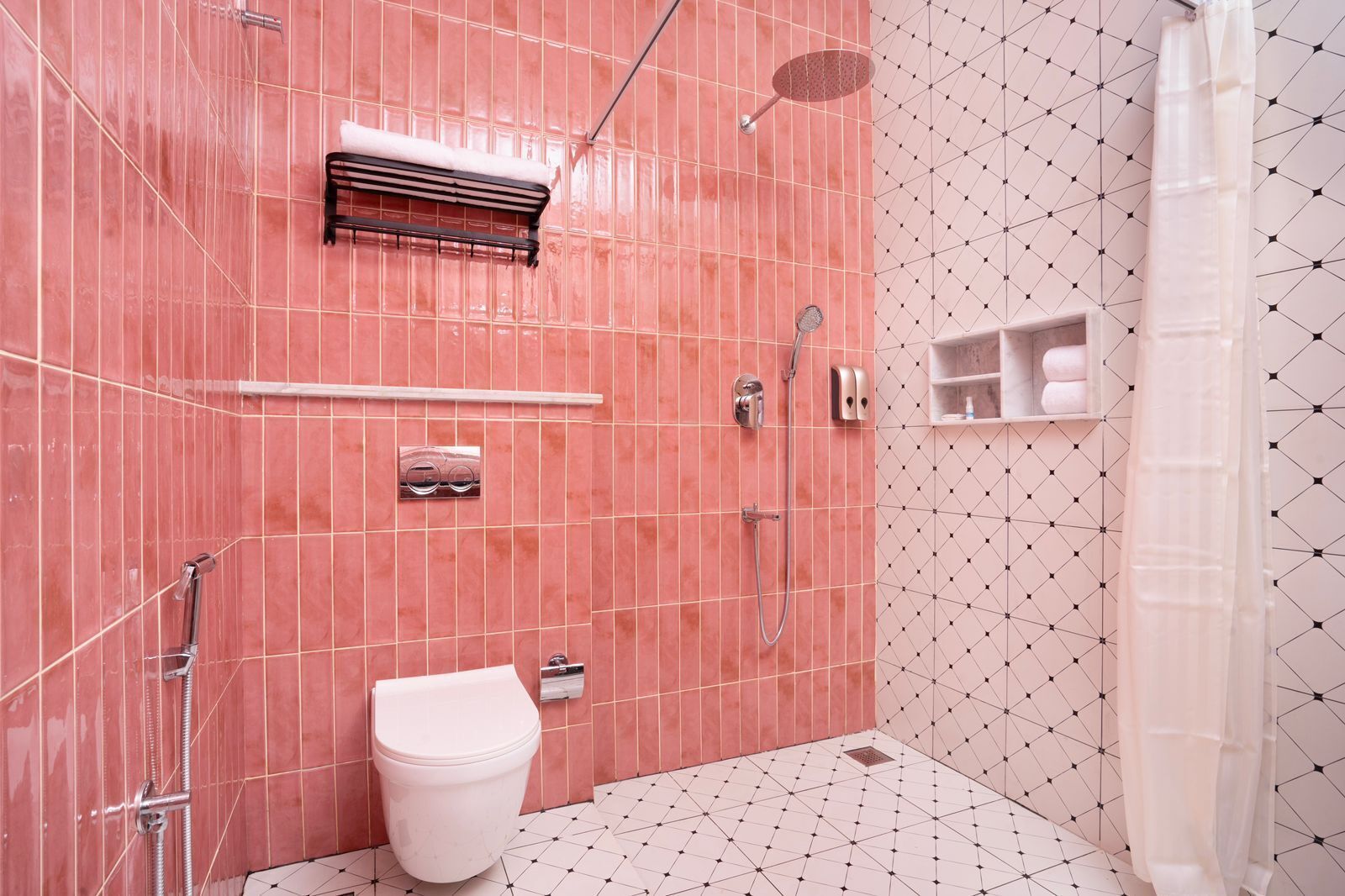 Baño con pared de azulejos rosa, inodoro blanco, zona de ducha y estante con toallas.