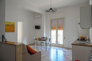A modern kitchen with a dining area, a TV mounted on the wall, and a large window with blinds.