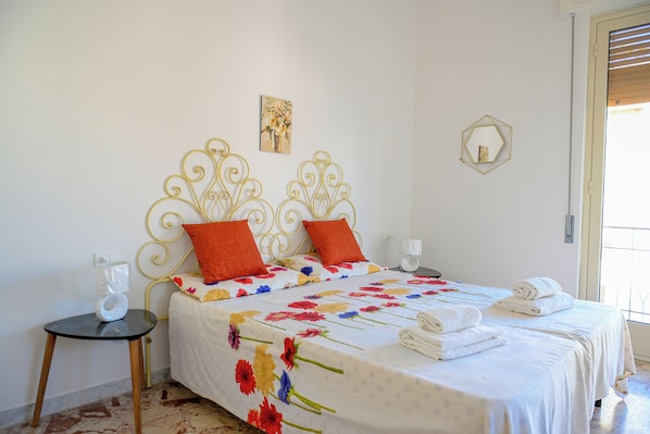 Une chambre à coucher avec une couvre-lit à motifs floraux, une tête de lit dorée et décorée, deux oreillers, une table de chevet avec une lampe et une fenêtre avec des stores.