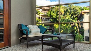 Un canapé en osier agrémenté de coussins et une petite table dans une pièce dotée d’une grande fenêtre donnant sur un jardin luxuriant et un immeuble avec des balcons.