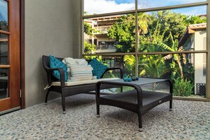 A wicker sofa with cushions and a small table in a room with a large window overlooking a lush garden and a building with balconies.