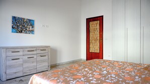 A bedroom with a bed, a wooden dresser, a red door, and a wall-mounted painting.