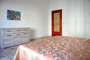 A bedroom with a bed, a wooden dresser, a red door, and a wall-mounted painting.