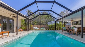A covered swimming pool with a glass ceiling, wooden deck chairs, and a patio area with potted plants.