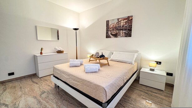 A modern hotel room with a large bed, a bedside table with a lamp, a nightstand, a dresser, a mirror, and a painting on the wall. - Casa Micio - MS 3 NT by Ca-sa Check (Chioggia)