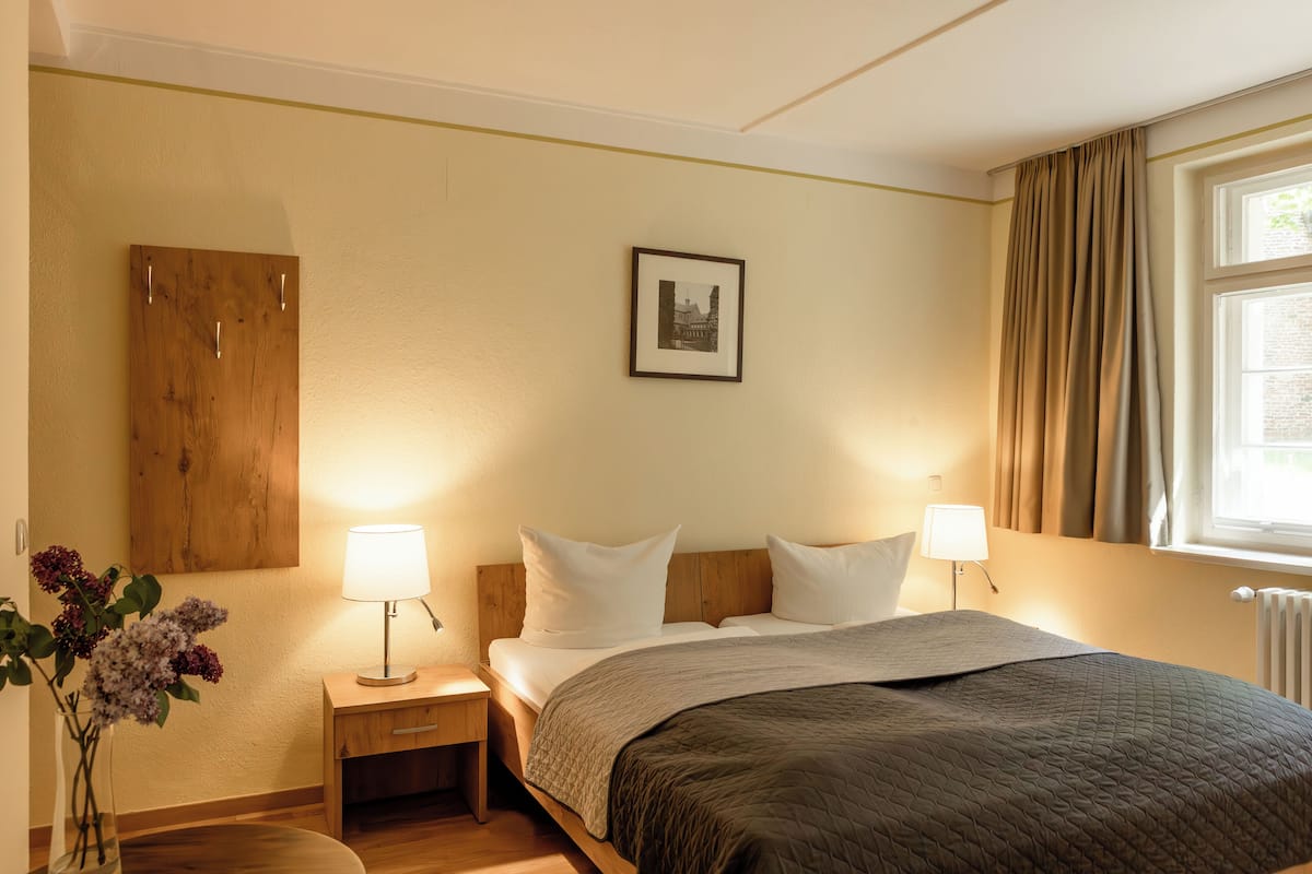 Une chambre d’hôtel avec un lit, des lampes de chevet, une table de nuit en bois, un tableau encadré au mur et une fenêtre avec des rideaux.