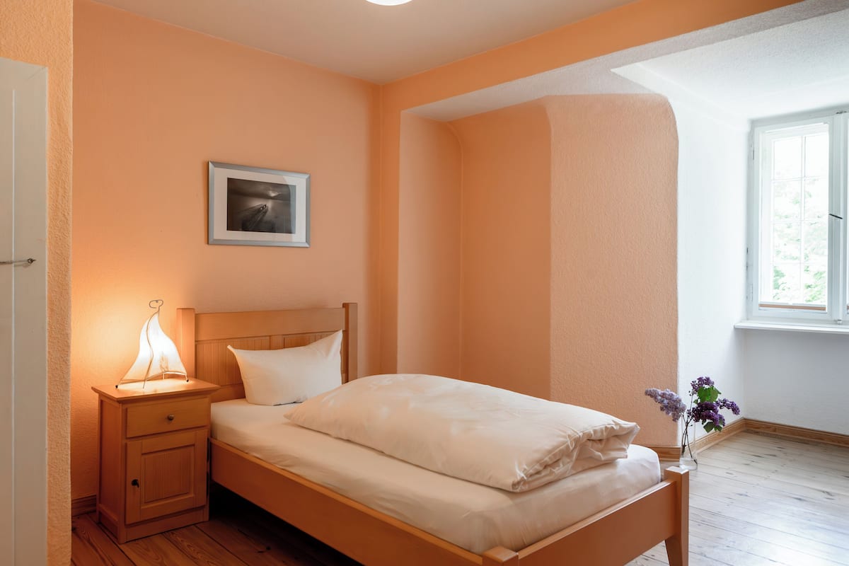Une chambre avec un lit en bois, une table de chevet avec une lampe, un tableau encadré au mur et une fenêtre donnant sur un paysage verdoyant.