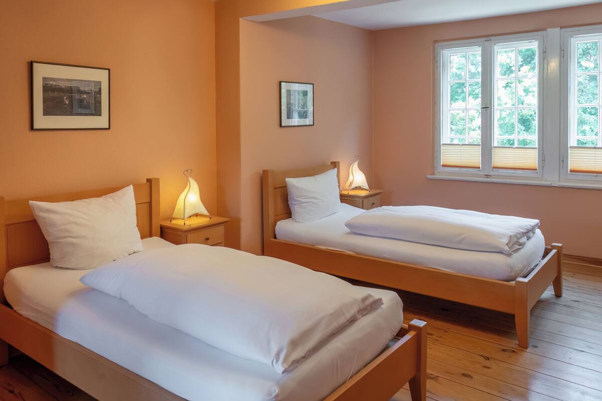 Une chambre avec deux lits simples, des têtes de lit en bois, des tables de chevet avec des lampes et une fenêtre avec des stores.