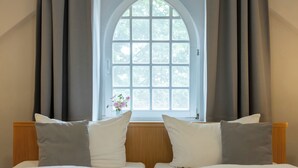 A bedroom with a large arched window, two beds with white and grey bedding, and a vase with flowers on the windowsill. - Klosterhotel Lehnin (Kloster Lehnin)