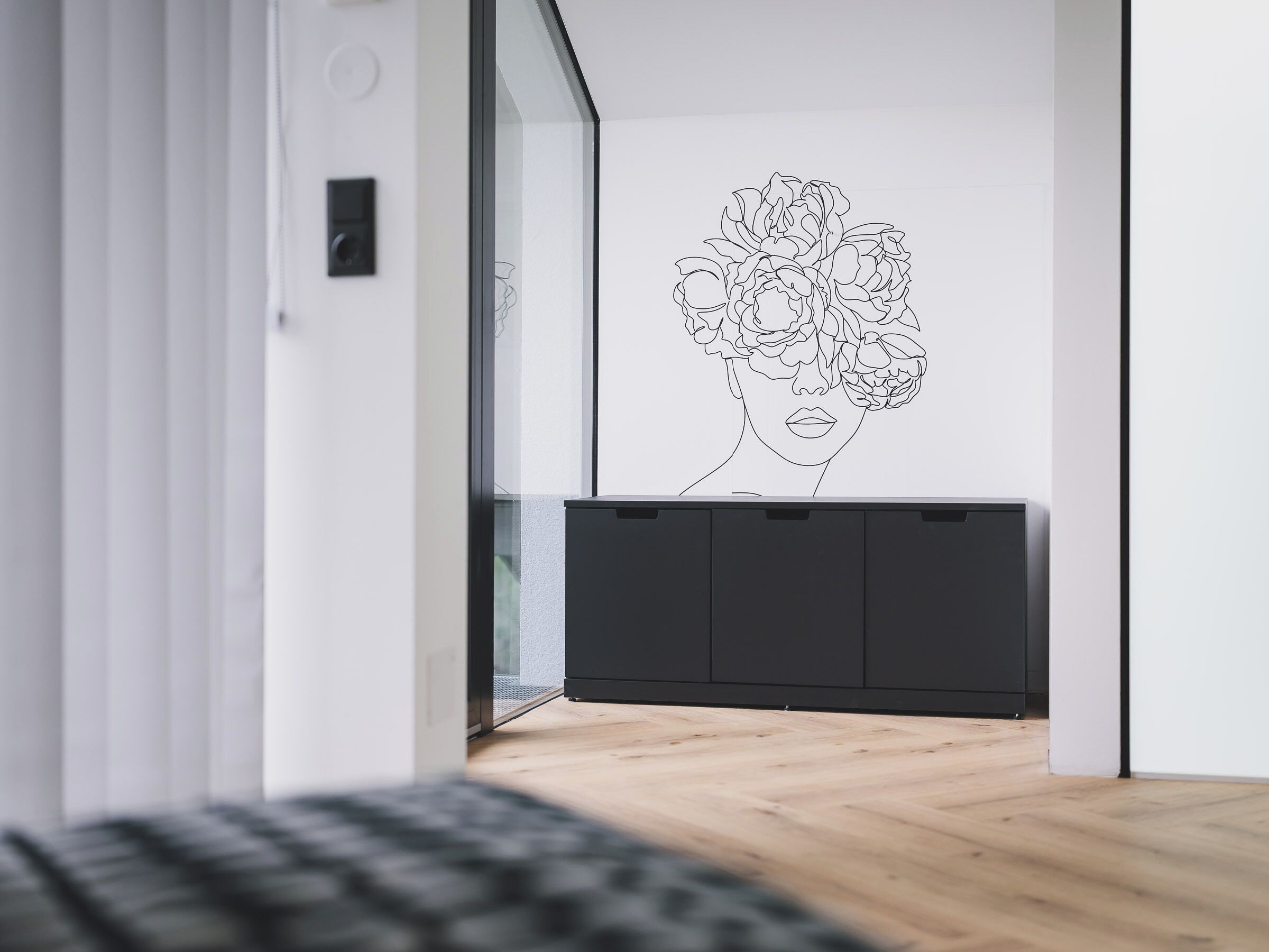 Un interior moderno con diseño minimalista, que presenta un piso de madera, una consola negra y una obra de arte floral en la pared.