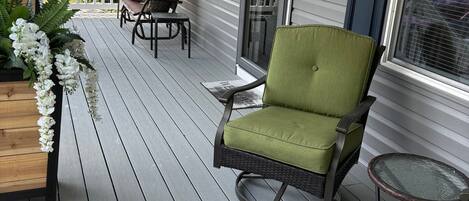 A wooden deck with a green chair and a small table, a white railing, and a window with shutters.