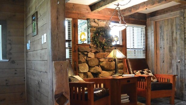 A rustic cabin interior with wooden walls, a stone fireplace, a wooden desk with a lamp, and a leather chair. - Spacious Rural Farmhouse Getaway with Peaceful Views (Williamsville)