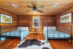 Une cabane avec deux lits en métal, des murs en bois, un ventilateur de plafond et une grande fenêtre.