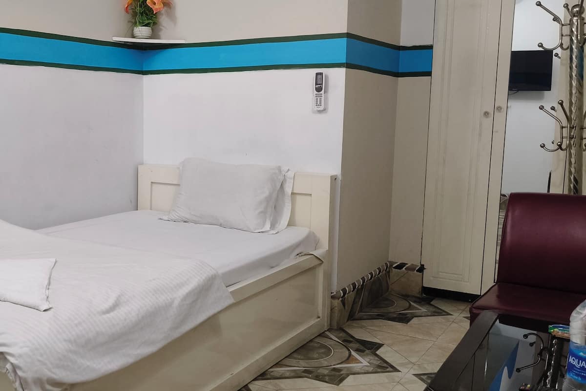 Una cama individual con colchón y almohada blancos, una mesita de noche pequeña, una silla, un teléfono de pared y un cubo de basura.
