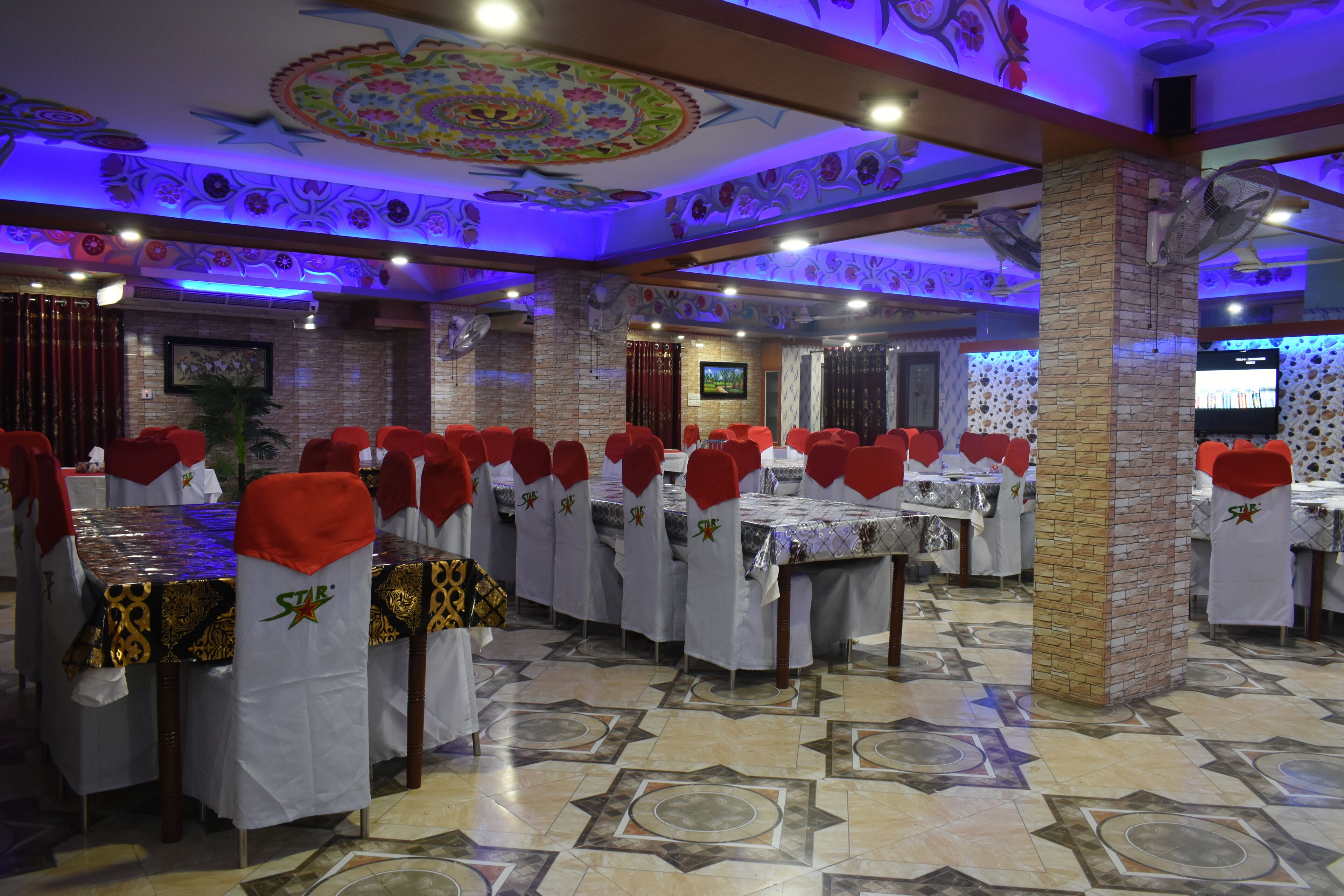 A restaurant with multiple tables and chairs, each with a red chair cover and a white tablecloth. The ceiling features intricate floral designs, and the floor has a patterned tile design.