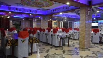 A restaurant with multiple tables and chairs, each with a red chair cover and a white tablecloth. The ceiling features intricate floral designs, and the floor has a patterned tile design.