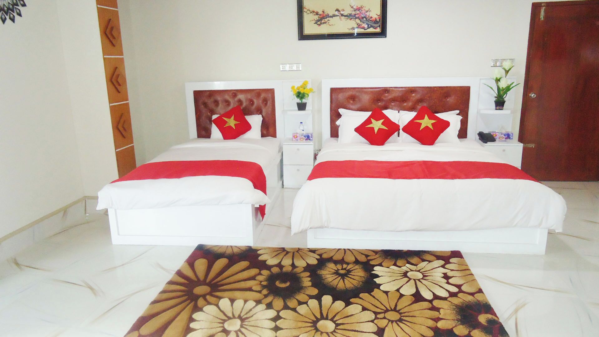 Habitación de hotel con dos camas, cada una con una colcha roja y blanca y almohadas estampadas con motivos de estrellas. Hay una mesita de noche con un jarrón de flores y una botella de agua. El piso es de baldosas y hay un cuadro en la pared.